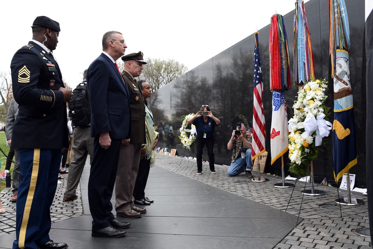 Three individuals stand near each other facing a wreath. The wreath is positioned near a black wall and an array of flags.