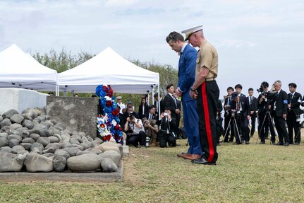 Defense Secretary Pete Hegseth and the Marine Corps commandant stand with bowed heads in front of a rock memorial with people and tents in the background.