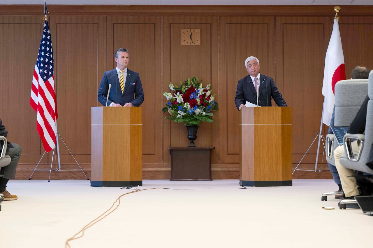 Two men speak from podiums in front of reporters.