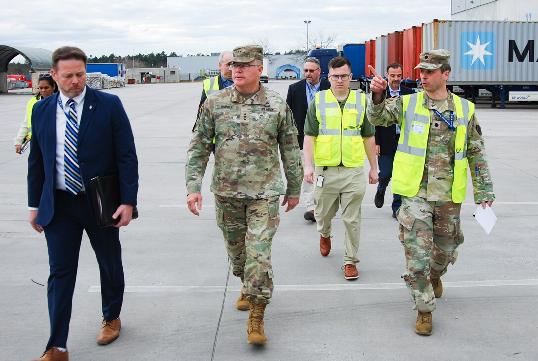 A group of people, including two men wearing military camouflage uniforms, walk outside a distribution center with shipping trucks in the background.