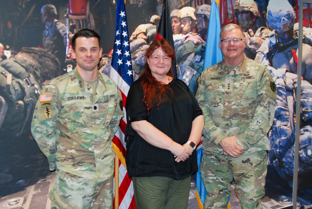 Two men in military camouflage uniforms stand on either side of a woman as they all pose for a photo.