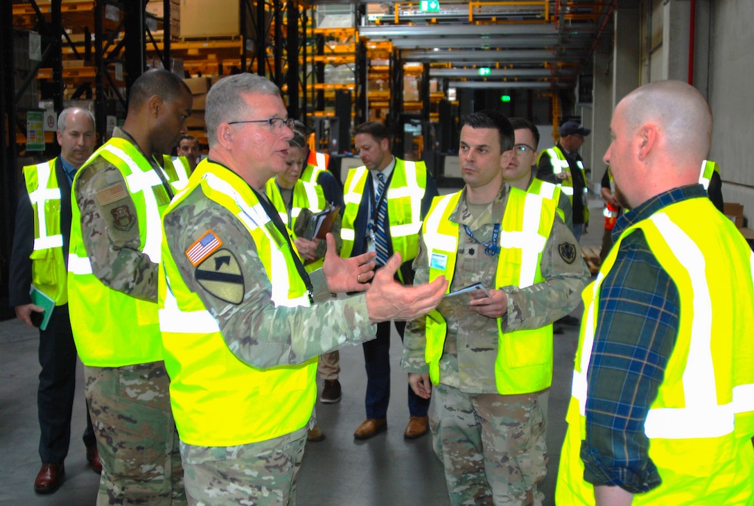 A large group of people wearing reflective safety vests stand in a warehouse having a discussion.