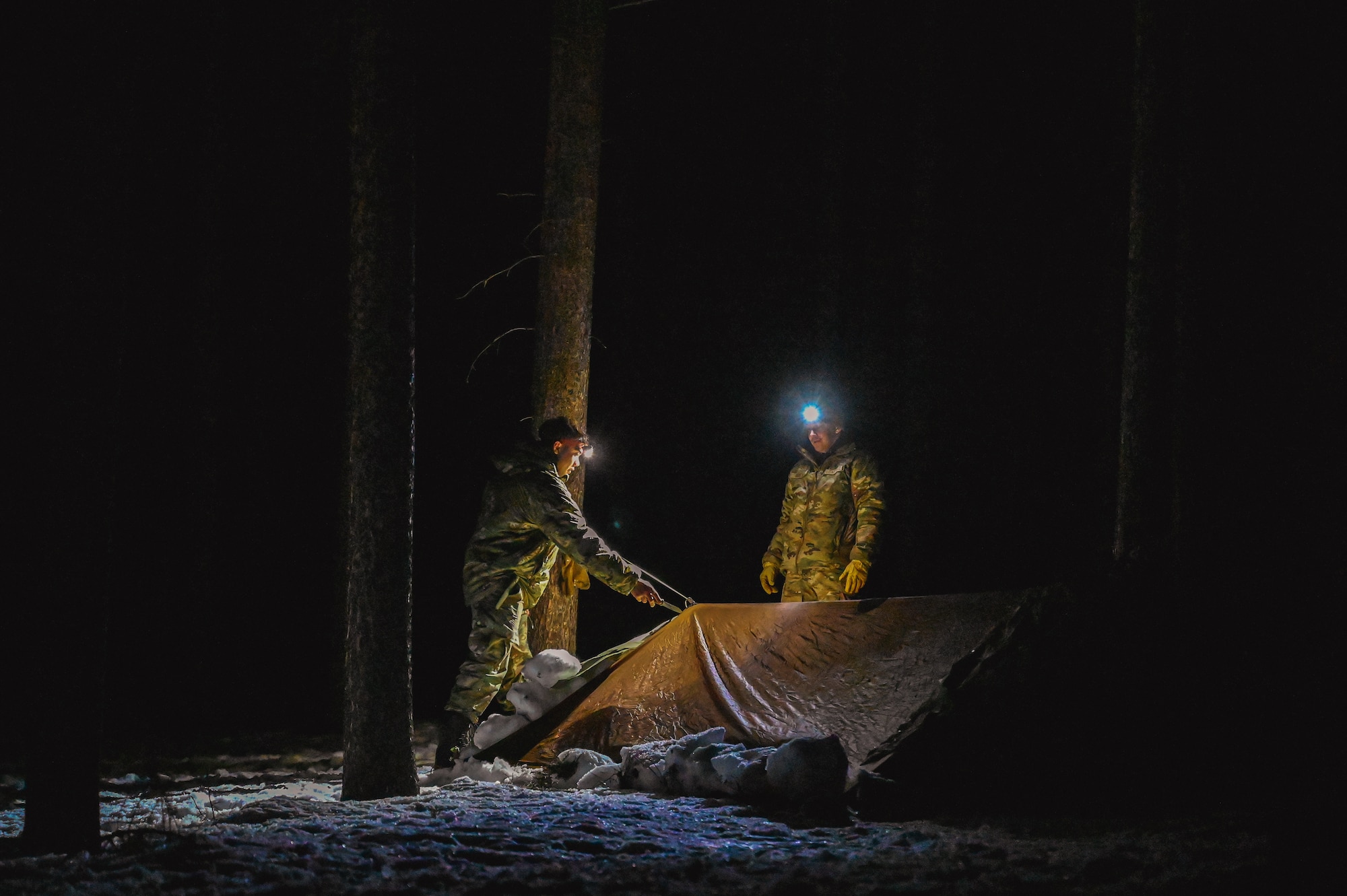 Two Airmen with headlamps stand next to a tent surrounded by darkness