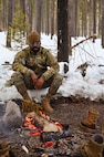 Airman sits in snow next to a fire in the woods