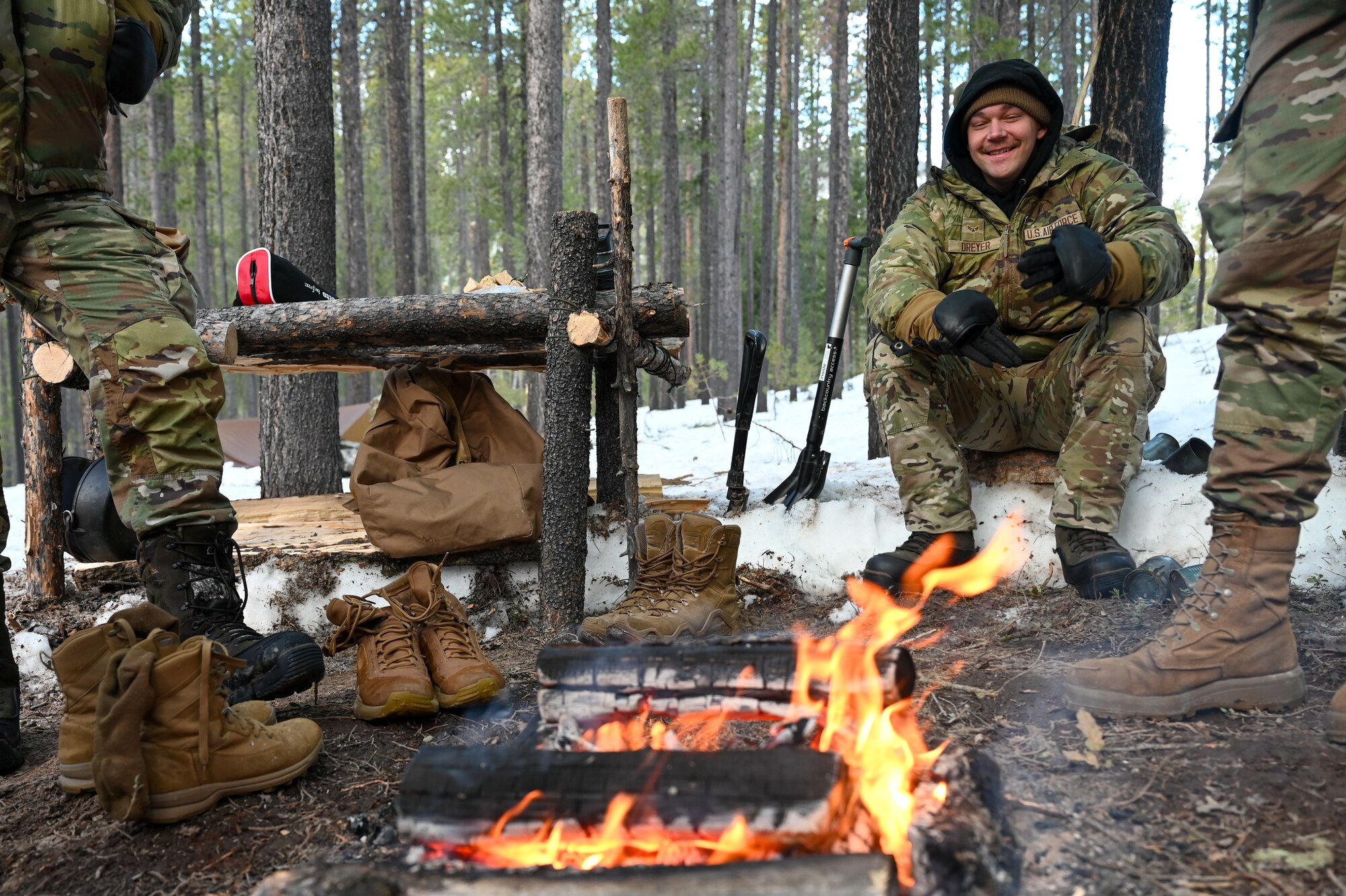 Airmen in uniform gather around a fire in the woods