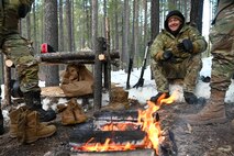 Airmen in uniform gather around a fire in the woods