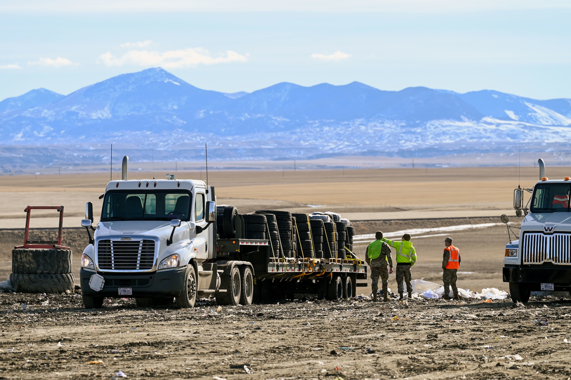 Three Airmen in high visibility vests stand next to a forty-foot flatbed truck loaded with pallets of tires with mountains in the background