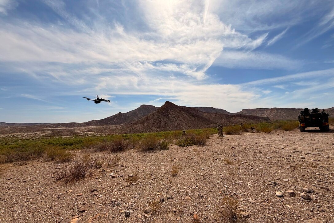 A drone flies above soldiers in a sparse field on a sunny day.