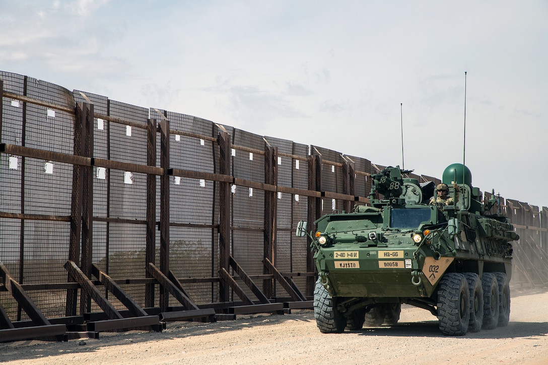 An armored military vehicle drives on a dusty road next to a tall fence.