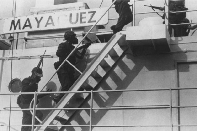 Military personnel holding weapons climb a ship’s ladder.