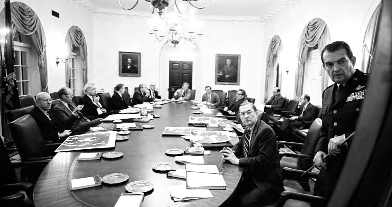 A man holding a stick briefs people seated at a table.