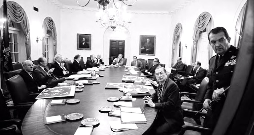 A man holding a stick briefs people seated at a table.