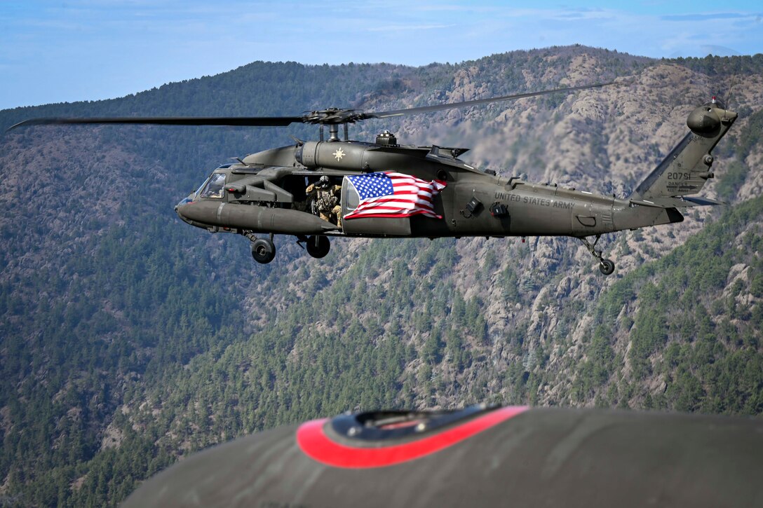 A soldier salutes out of a helicopter with an American flag over a mountainous forest.