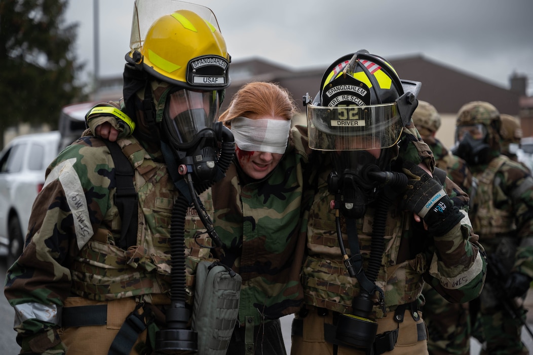 Military firefighters in hazmat suits and combat kit carry a patient with a bandage over her eyes.