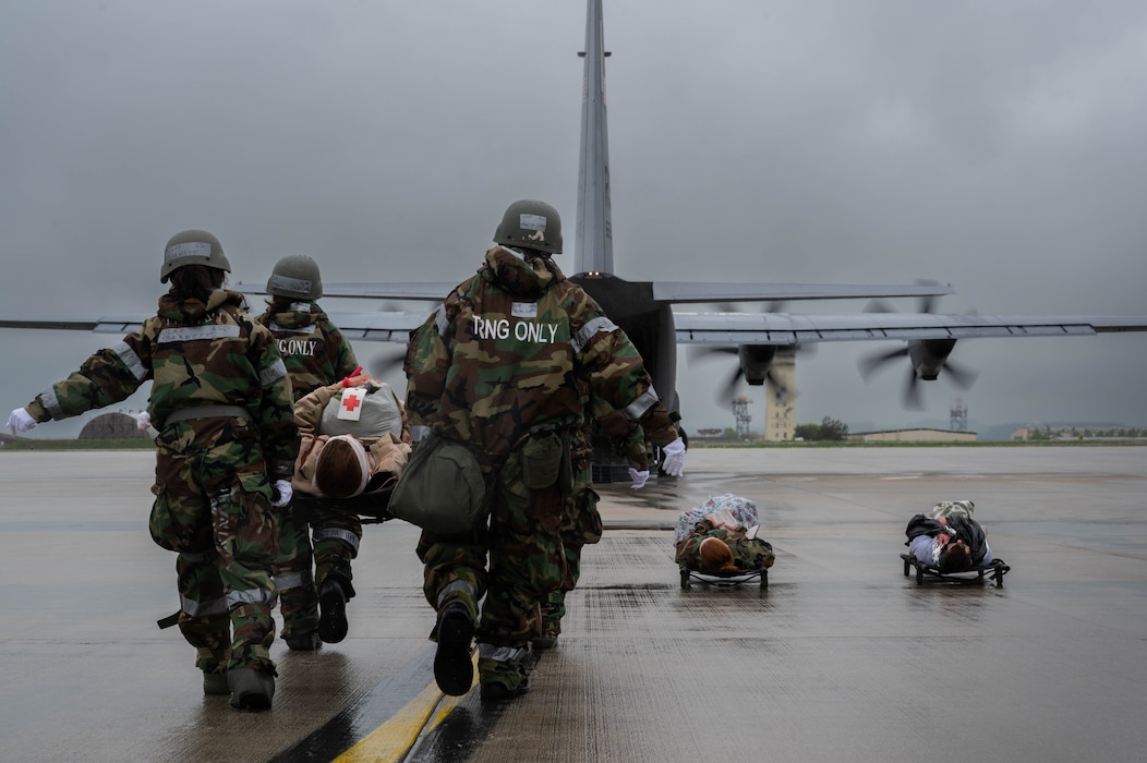 Military medics wearing hazmat suits and combat kit carry a patient on a litter towards a C-130 transportation aircraft.