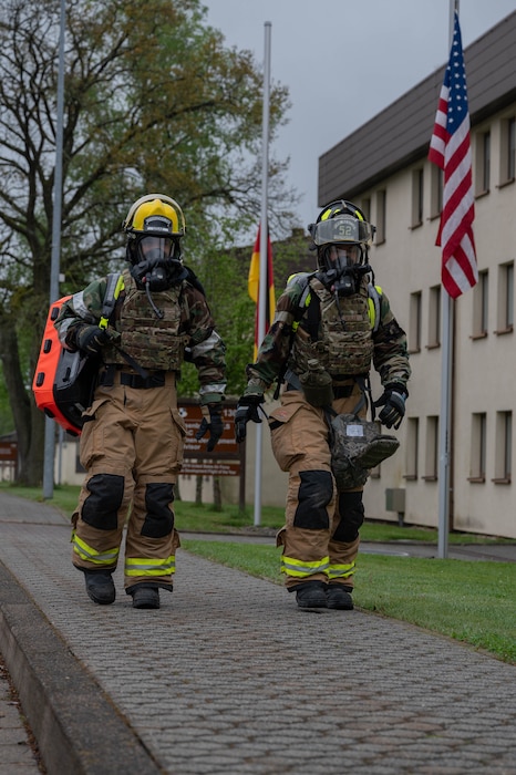 Military firefighters wearing hazmat suits and protective gear carry medical equipment down a road.