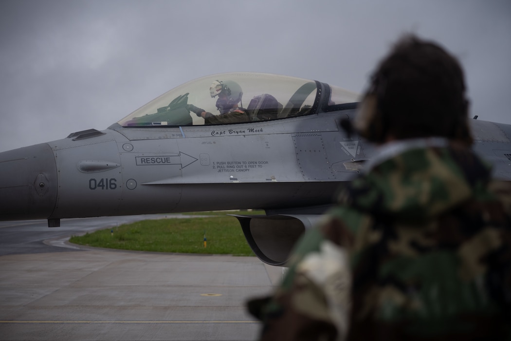 A mechanic wearing a hazmat suit watches as a fighter pilot taxis his fighter jet.