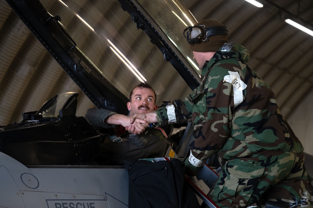 A fighter pilot sitting in a cockpit shakes hands with a mechanic wearing a hazmat suit.