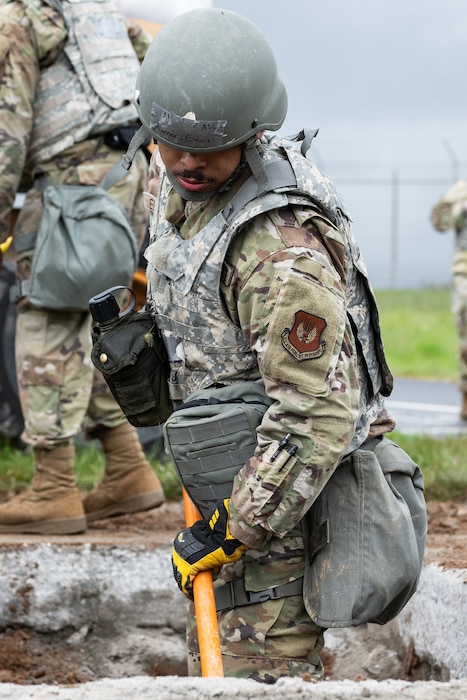 An Airman in full combat kit stands in and shovels in a square hole on a taxiway.
