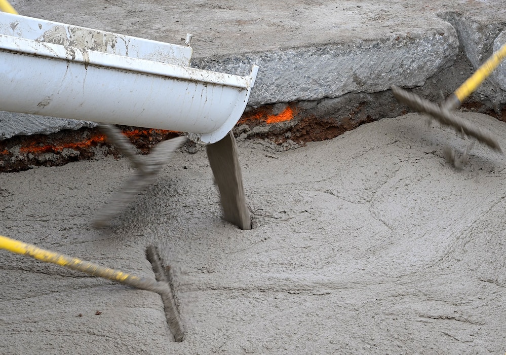 Concrete pours out of a white trough into a hole in a taxiway while smoothing tools spread it throughout the hole.