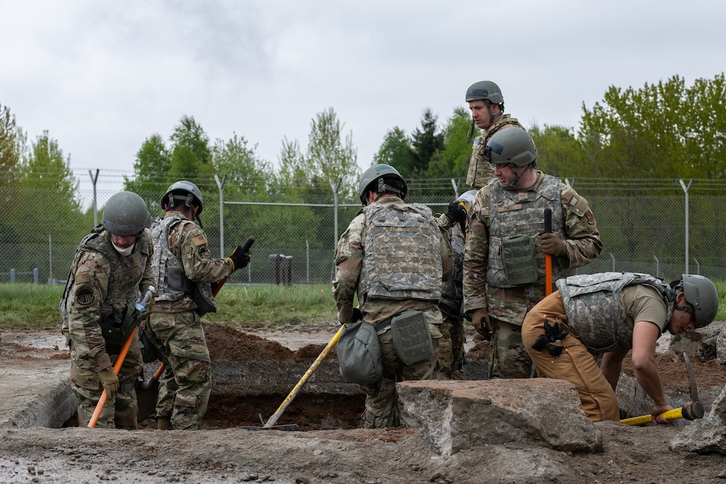 Airmen in full combat kit stand in and shovel in a square hold surrounded by concrete and dirt debris on a flightline.