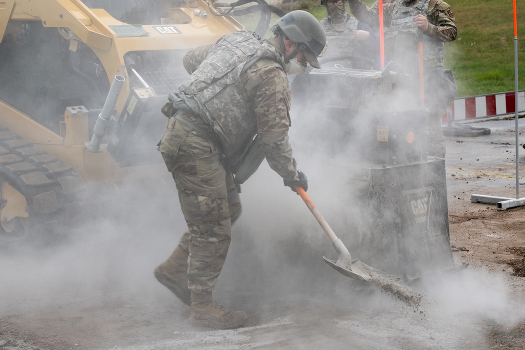 An Airman in full combat kit shovels powdered concrete away from a giant saw cutting concrete while surrounded by grey dust.