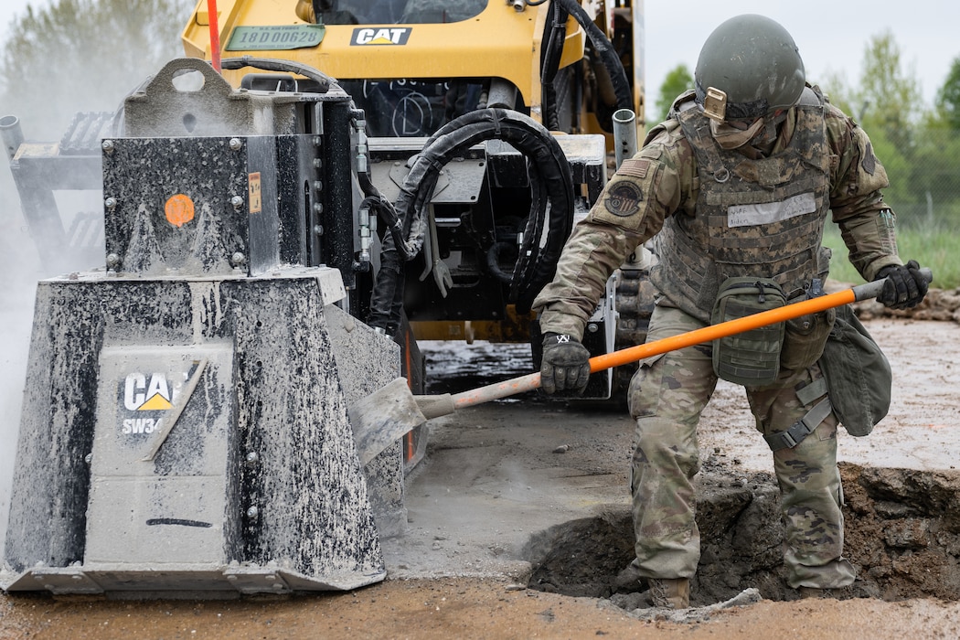 An Airman in full combat kit shovels powdered concrete away from a giant saw cutting concrete while standing in a crater.