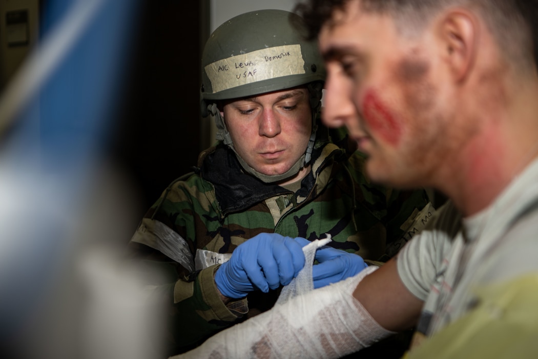 A military medic in a hazmat suit and helmet wraps a patient's arm in a bandage.