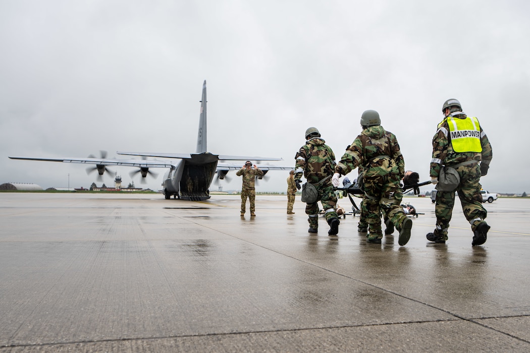 Four military medics in hazmat suits carry a patient on a litter towards a C-130 transport aircraft.