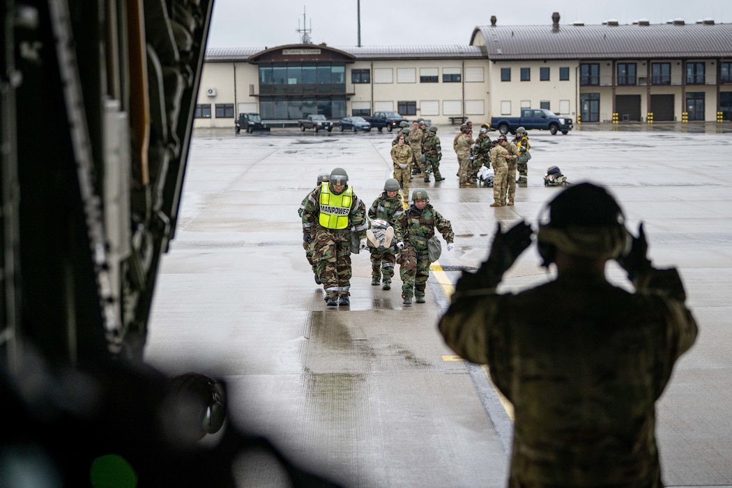 A C-130 loadmasters motions a team of four military medics in hazmat suits to carry a patient on a litter into the aircraft.