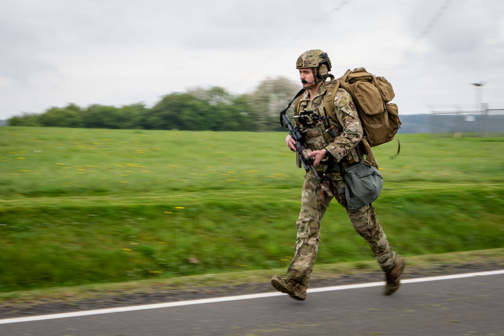 A military policeman wearing protective gear and a ruck sack runs down a road while carrying a weapon.