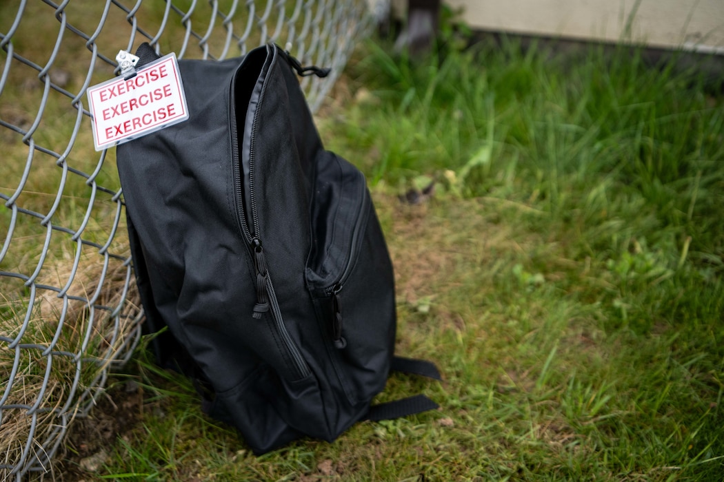 A black backpack with a tag that says EXERCISE lies up against a chain-link fence.