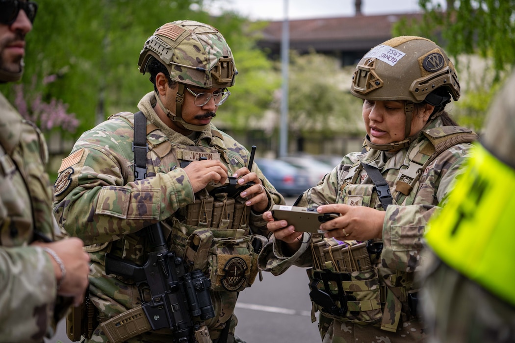 Airmen in vests and helmets look at a phone.