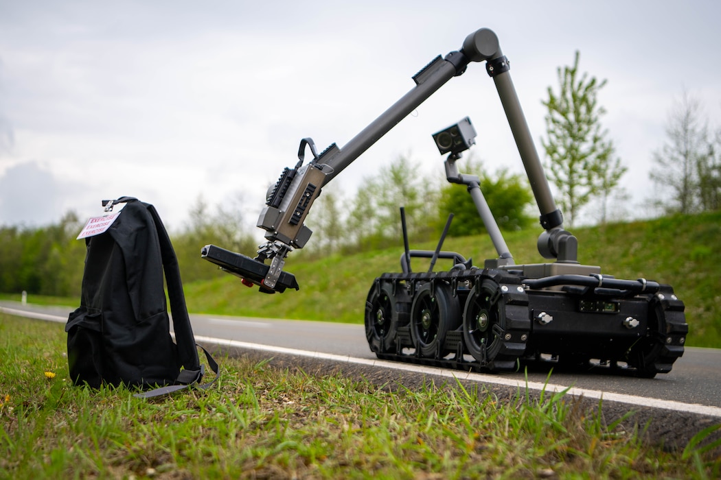 A small, tracked robot sticks its robotic arm at a black backpack.
