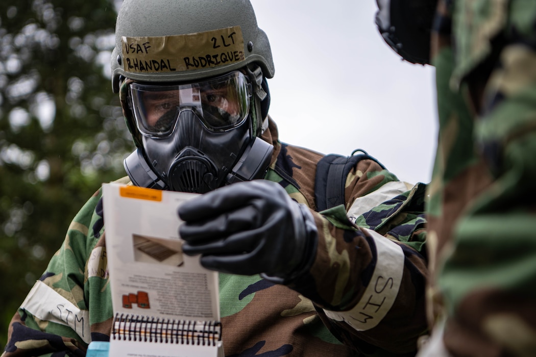 An Airman wearing a chemsuit, gas mask and helmet reviews a quick reference notebook.