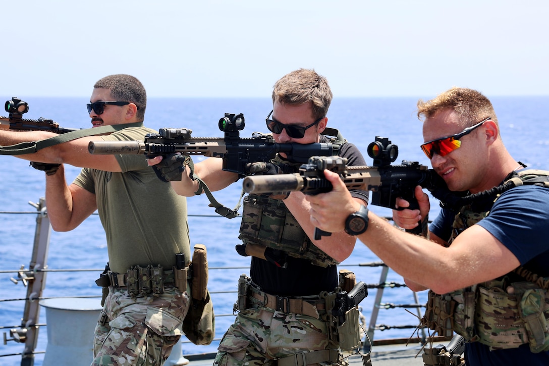 Three Coast Guardsmen aim weapons aboard a ship at sea.