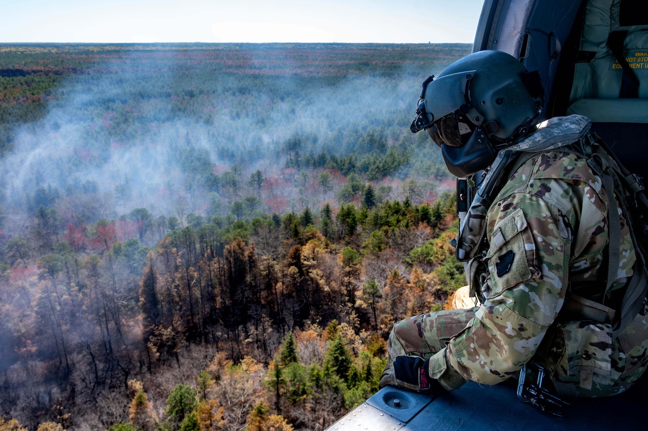 A guardsman watches from the cabin of an open helicopter into a smoky forest below.