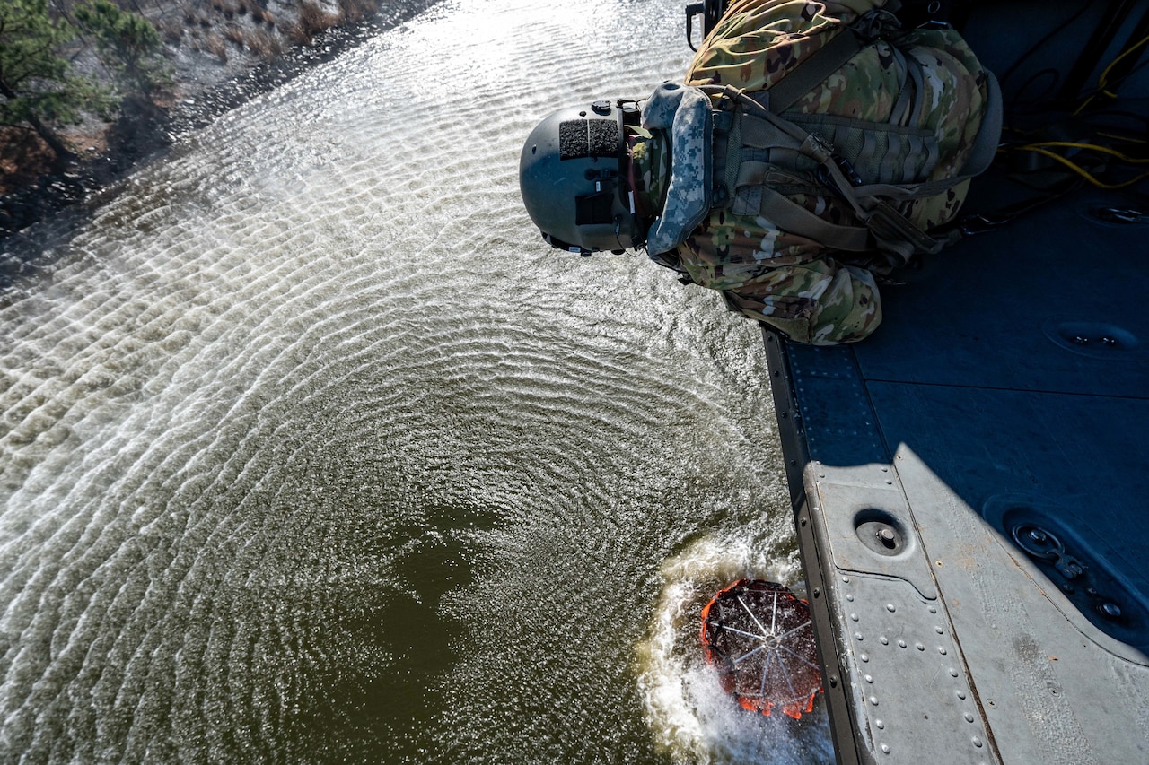 An airman leans over the open cabin of a helicopter as a bucket is filled with river water below.