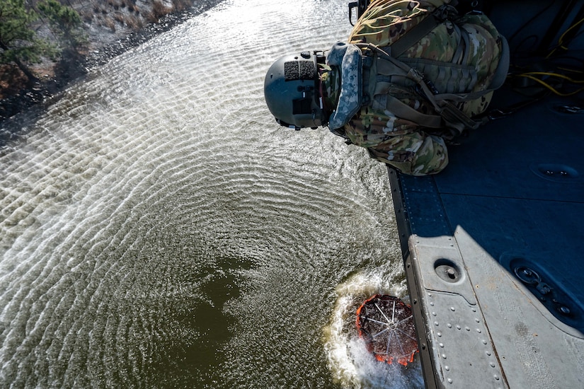 An airman leans over the open cabin of a helicopter as a bucket is filled with river water below.