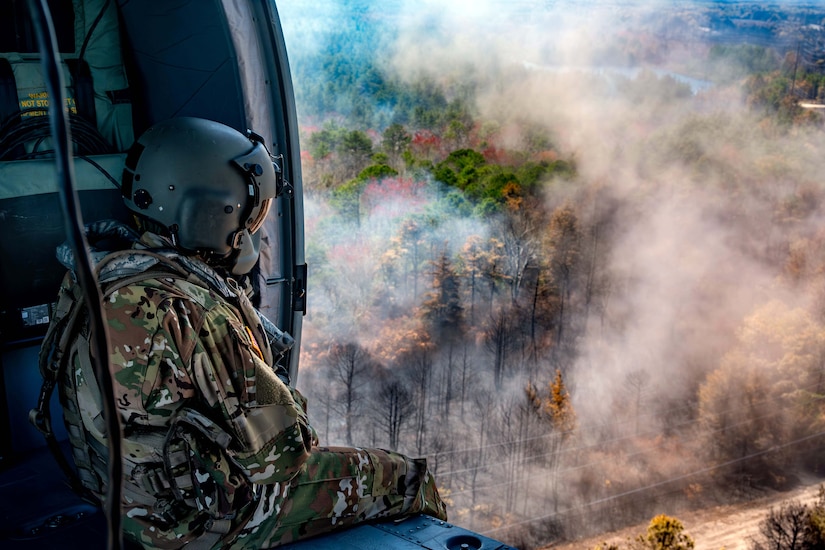 An airman sits in a helicopter's open cabin watching a smoky forest below.