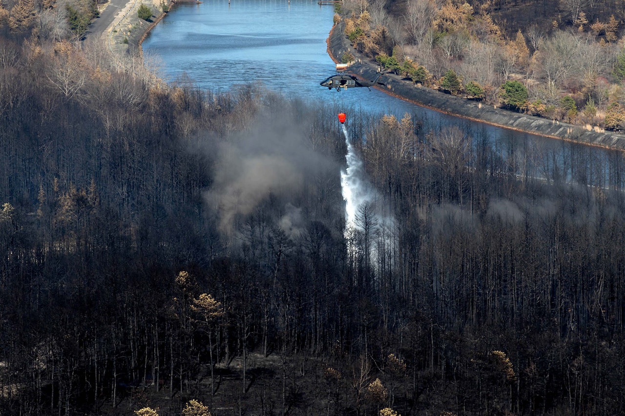 A helicopter drops water from a red bucket over scorched trees, with smoke billowing.