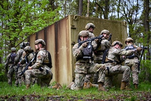 Airmen in tactical gear face left while another group faces right against a rusted container in a forest setting.