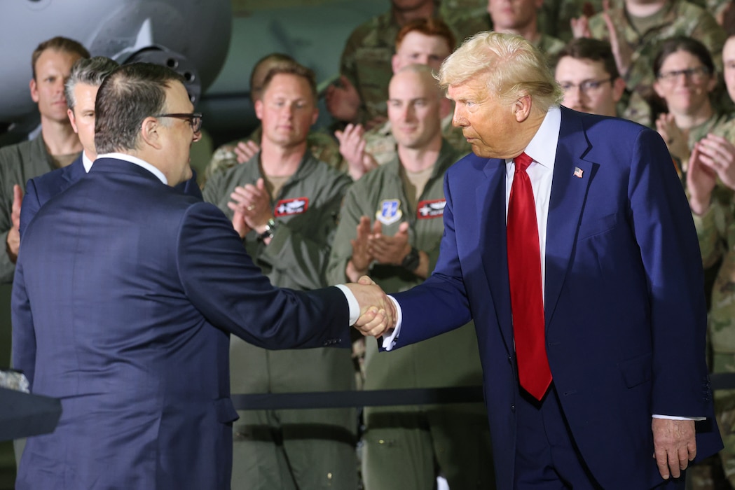 Two men in civilian clothes shake hands with servicemembers standing and clapping behind.