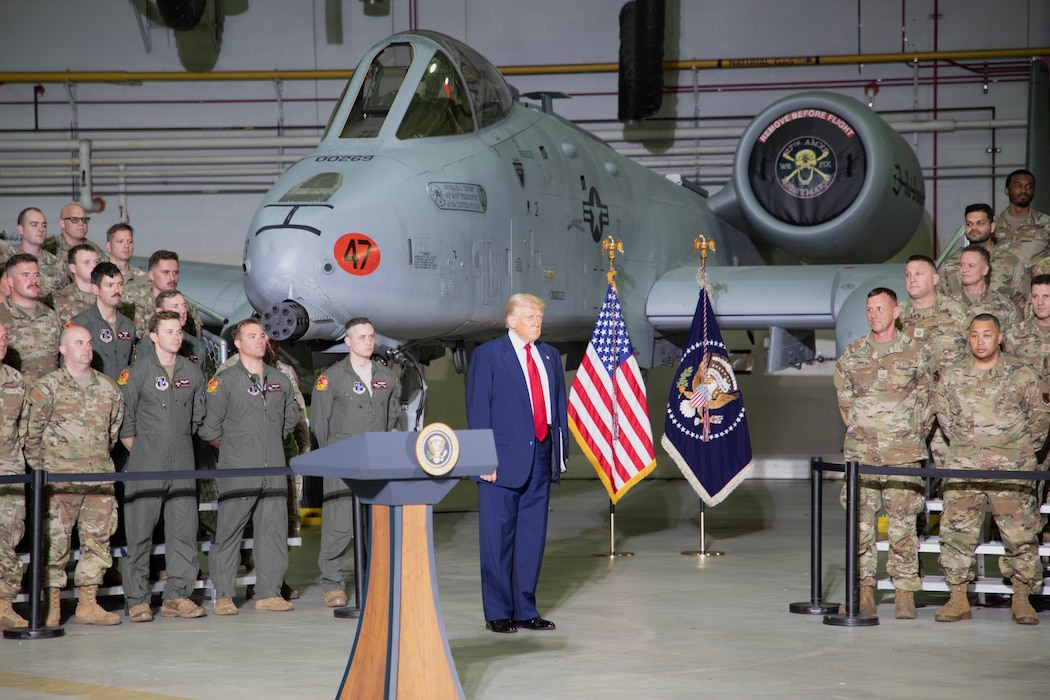 President stands in hangar next to U.S. flag, while servicemembers in uniform stand in audience.
