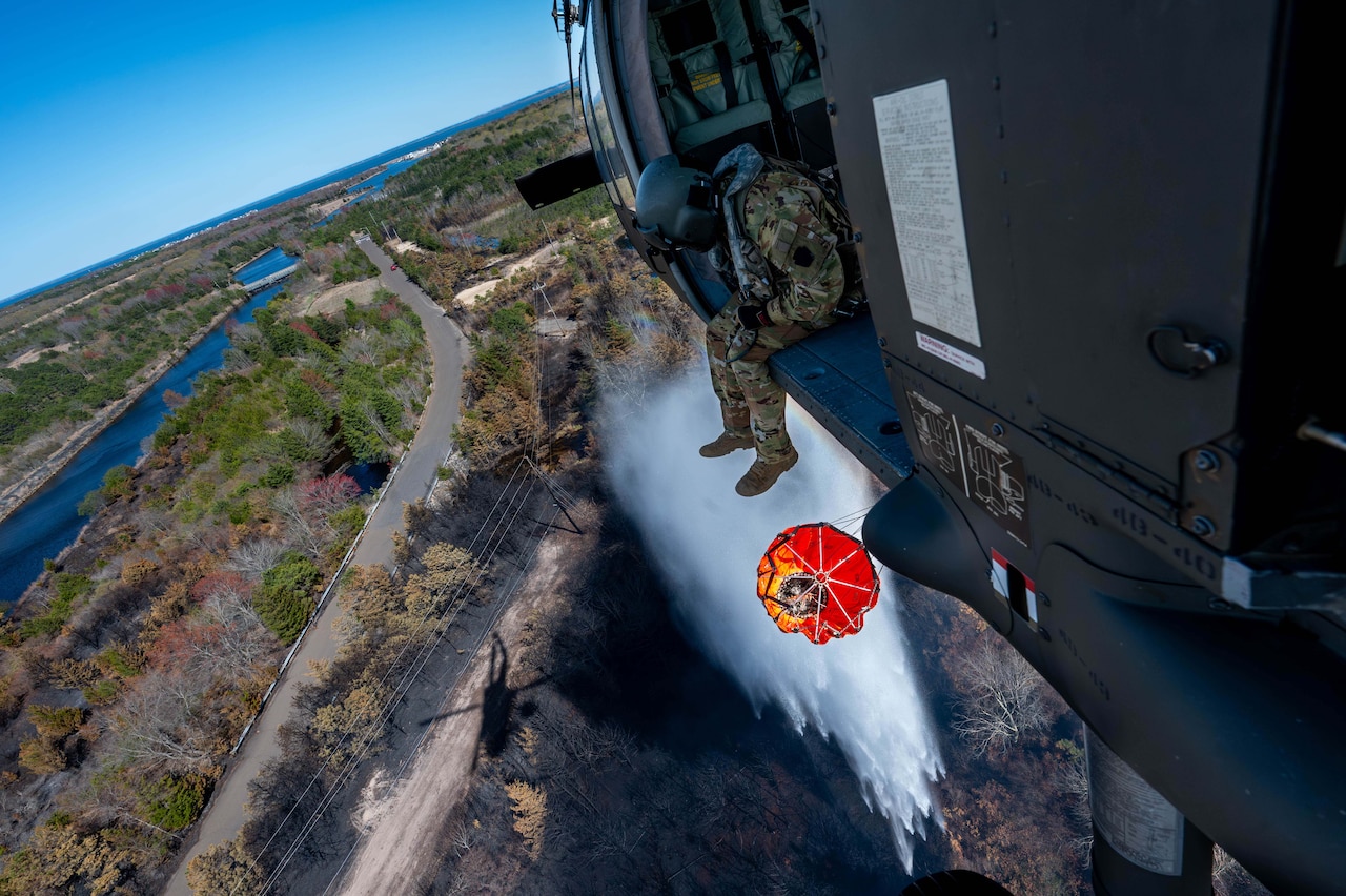 A soldier sits in a helicopter and observes an orange firefighting bucket drop water on a smoky forest.