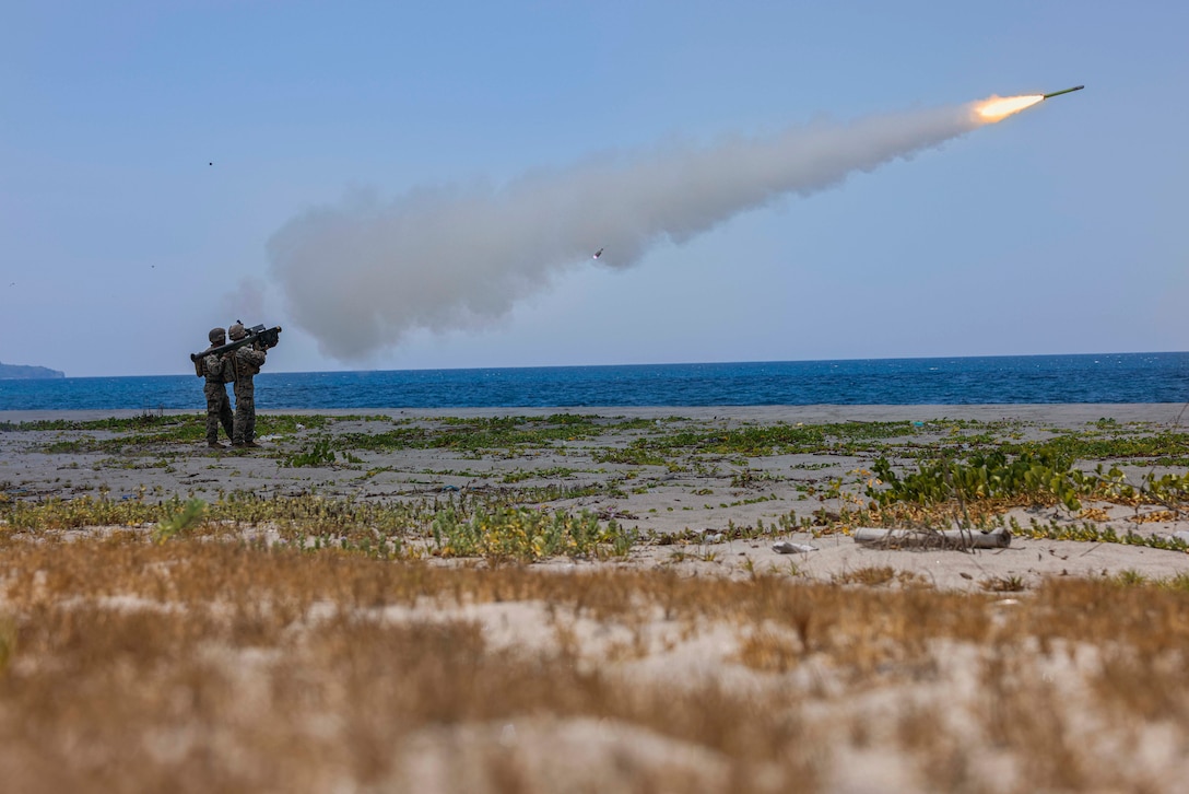 Two Marines on a beach fire a missile towards the ocean, leaving behind smoke.