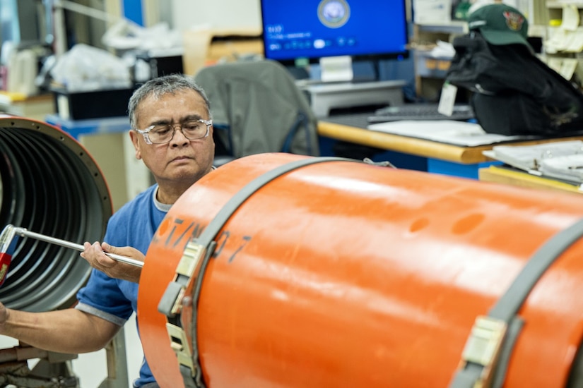 A man in a blue shirt holding a tool looks into the depths of a cylindrical fuel tank.