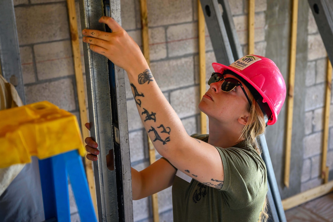A Navy hospital corpsman wearing a construction helmet holds a leveling tool in a building being renovated.
