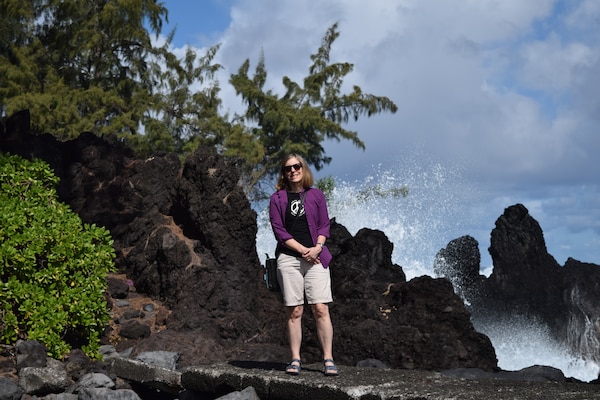 A woman in a purple shirt and tan shorts stands on rocks with waves splashing in the background.