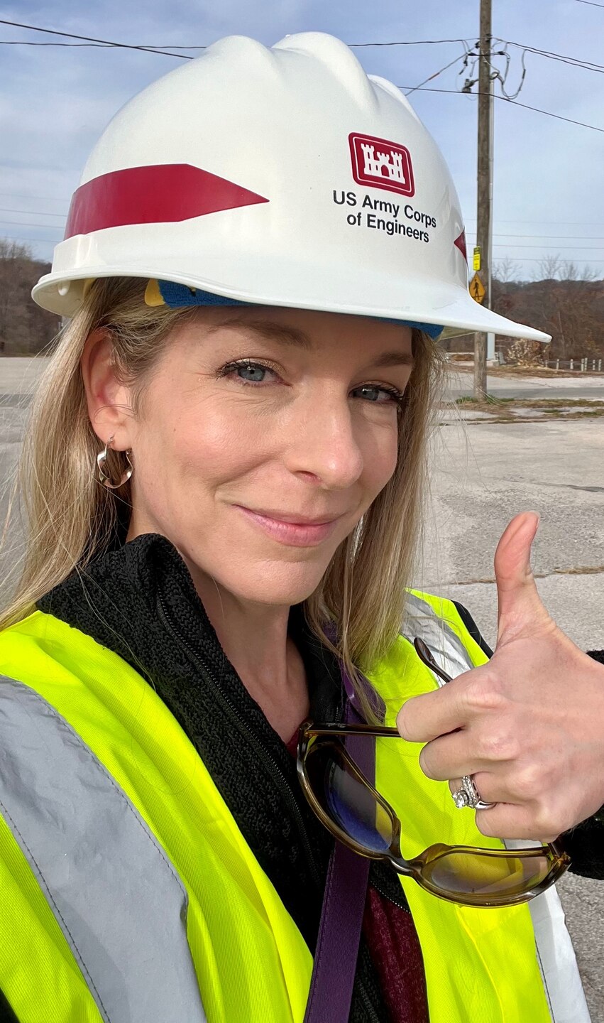 A woman in a white hardhat and a yellow safety vest gives a thumbs up.